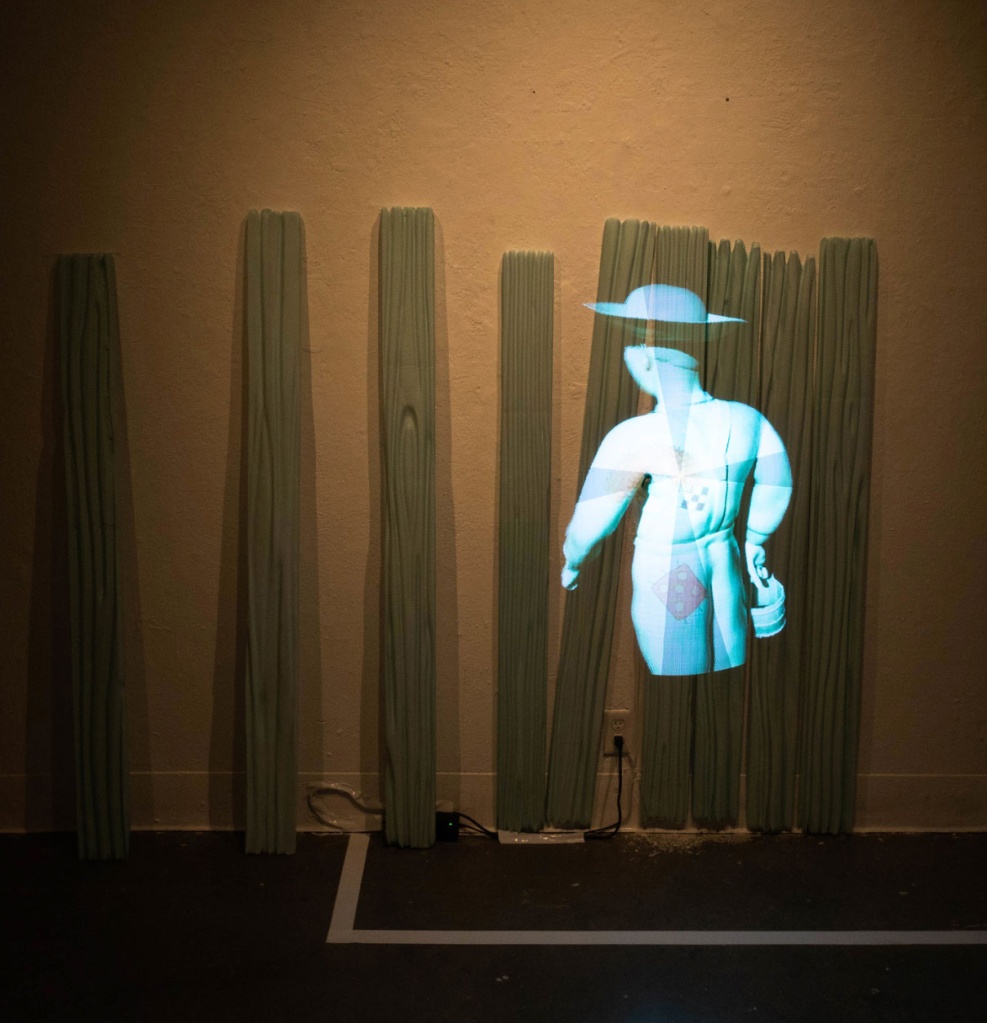 A projection of a figure wearing a broad hat on weathered fencing against a gallery wall